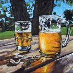 Two glasses of golden beer on picnic table in the shade of trees, blue skies and green lawn in the background